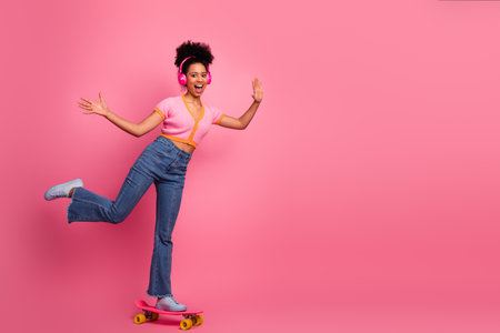 Happy young woman enjoying music while balancing joyfully on a skateboard against a vibrant pink...