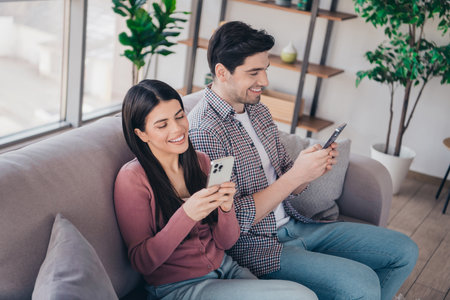 Young couple enjoying leisure time together at home, using smartphones while sitting on a cozy couch in a stylish apartment living room.の写真素材