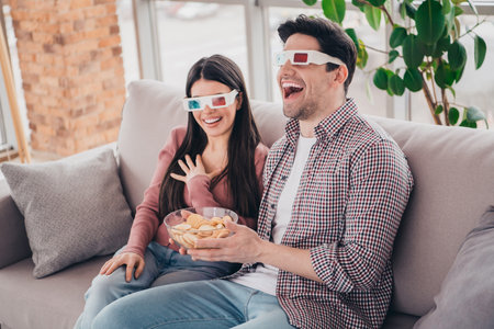 Happy young couple wearing 3D glasses enjoying snacks while watching a movie together at homeの写真素材