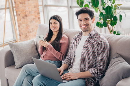 Happy couple enjoying leisure time together indoors, sitting on a sofa with a laptop and phone,...