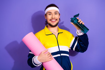 Happy man in retro sportswear holding a yoga mat and water bottle on a vibrant purple background, ready for fitnessの写真素材