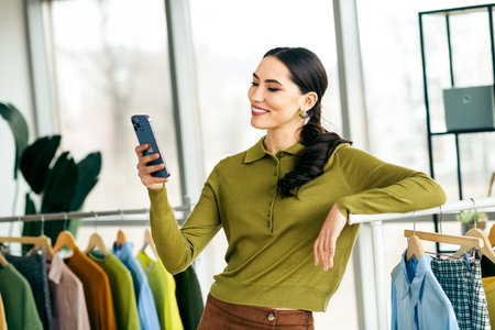 Confident female entrepreneur enjoying workplace productivity while using smartphone in a modern fashion studio settingの写真素材