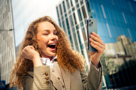 Excited young businesswoman holding smartphone on urban street, expressing success and happiness with vibrant emotionsの写真素材