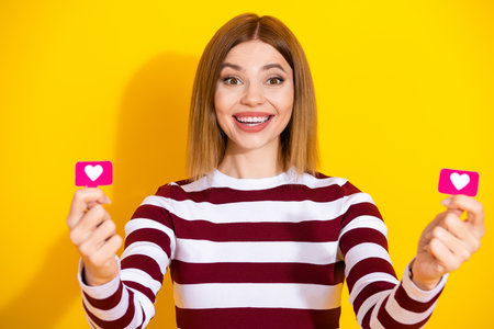 Young woman in striped sweater joyfully showing heart symbols against yellow background for creative...