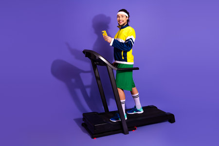 Retro-styled young athlete in bright activewear using smartphone on treadmill against colorful...