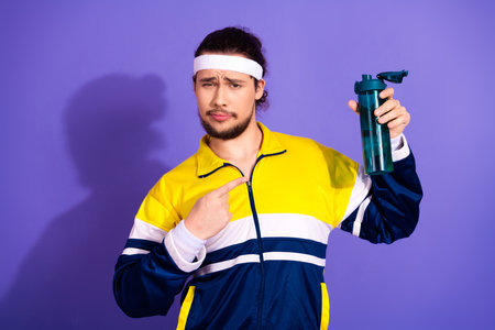 Confident young man in sportswear holding a bottle, pointing with a finger, against a vibrant purple...
