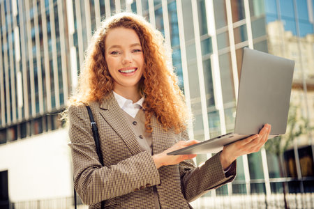 Young professional woman holding a laptop outdoors in an urban business district on a sunny dayの写真素材