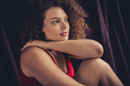 Confident young woman with curly brown hair and natural beauty in red outfit sitting gracefully against purple studio backgroundの写真素材