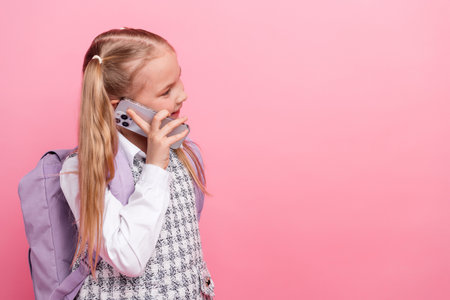 Smiling young schoolgirl talking on smartphone with cheerful expression, wearing uniform and backpack standing against pink backgroundの写真素材