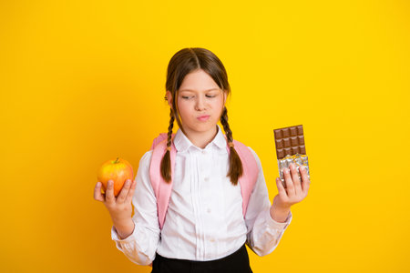 Young schoolgirl holds an apple and chocolate, weighing choices against a yellow background, symbolizing healthy decision-makingの写真素材