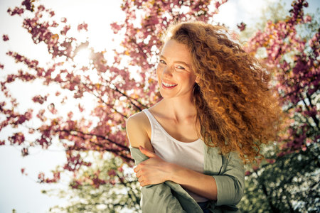 Charming young woman with curly red hair smiling in blooming park under daylight capturing spring sensationsの写真素材
