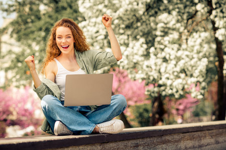 Lovely woman with red curly hair celebrating success with her laptop outdoors in a blooming park on a sunny dayの写真素材