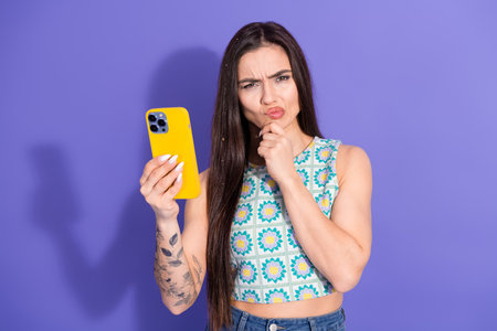 Young woman holding a yellow smartphone against a violet background with an expressive face and...