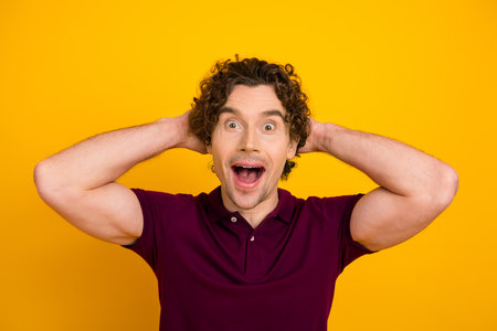 Excited young man with curly hair in a casual burgundy polo on a vibrant yellow background expressing surpriseの写真素材