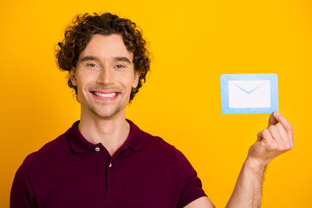 Smiling young man holding blue envelope icon card against yellow background symbolizing communication and positivityの写真素材