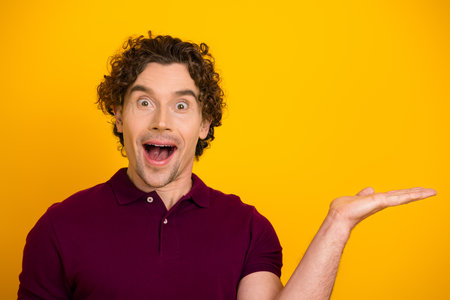 Surprised young man in burgundy polo gesturing energetically against bright yellow background with expressive excitementの写真素材
