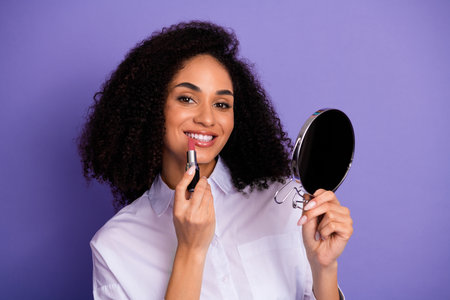Young woman with curly hair applying lipstick while holding a mirror, showcasing beauty, charm, and a confident smileの写真素材