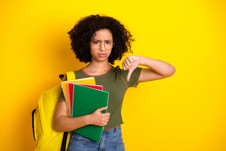 Young woman in casual attire showing a disapproving thumbs-down gesture, holding notebooksの写真素材