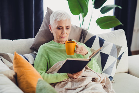Senior woman enjoying a relaxing day at home with a book and tea, surrounded by cozy pillows and a...
