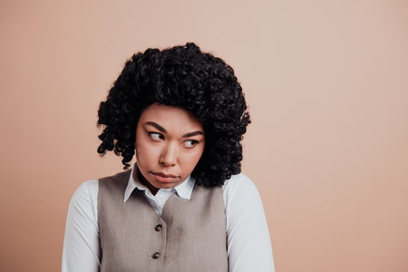Professional young woman in formal attire with expressive hairstyle looking thoughtful against beige...