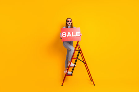 Young woman standing on a ladder holding a red sale sign against a bright yellow background...