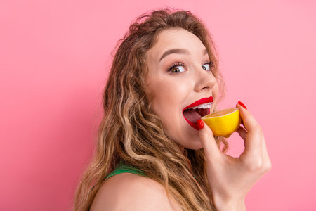 Portrait of a young woman holding a lemon and smiling against a vibrant pink background, expressing...