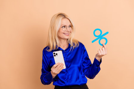Mature woman in formal blue shirt holding smartphone and percent symbol on beige background...