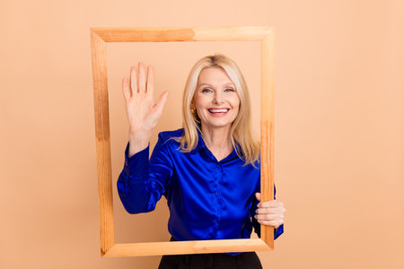 Elegant and cheerful mature woman gesturing with an open hand inside a wooden frame against a...
