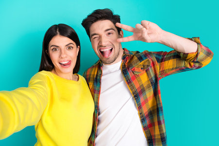 Happy young couple taking a selfie against a vibrant teal background showing cheerful gestures and stylish casual outfitsの写真素材