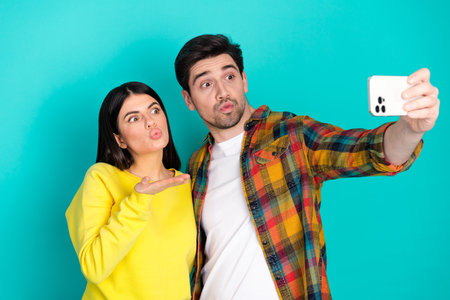 A young couple posing for a selfie with playful expressions against a teal background, enjoying a...
