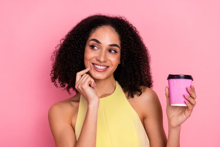 Smiling young woman with curly hair holding coffee cup against pink background, expressing happiness and lifestyleの写真素材