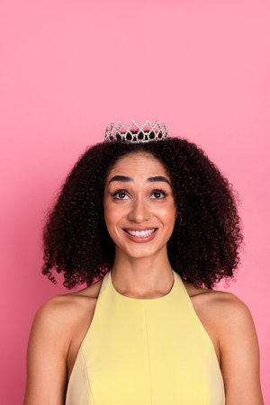 Young woman with natural curly hair wearing a yellow dress and a tiara smiles joyfully, standing against a pink backgroundの写真素材