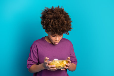 Young boy holding bowl of chips while standing against a vibrant blue background, wearing a purple...