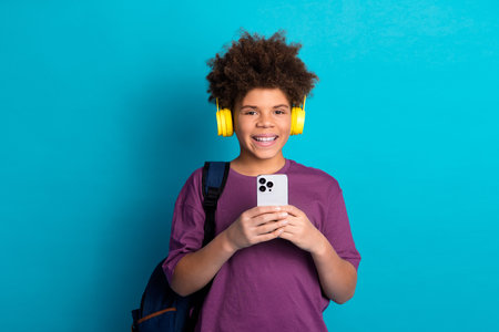Smiling teenage boy with curly hair enjoying music on headphones while holding a smartphone with a...