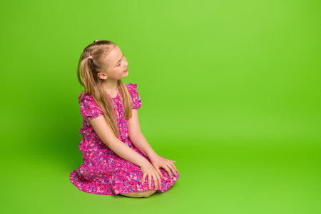 Portrait of a smiling young girl in a bright floral dress seated on a vibrant green background in a casual poseの写真素材