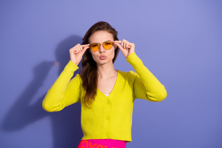 Stylish young woman in bright yellow cardigan posing with yellow sunglasses against vibrant purple backdropの写真素材