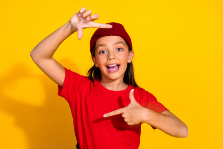 Excited young girl posing against yellow background, gesturing enthusiastically while wearing a red...