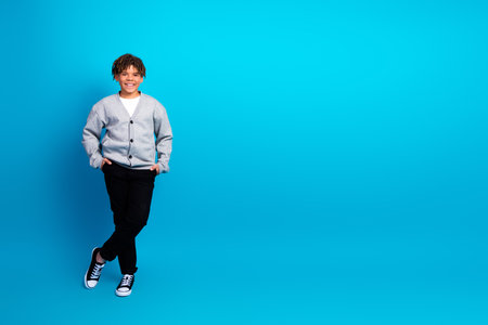 Smiling schoolboy wearing a light gray cardigan and black pants posing against a vivid blue backgroundの写真素材