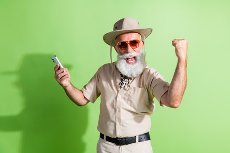 Photo of excited explorer senior man use online map cell phone raise winning fist isolated green...