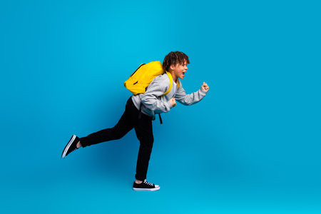 Energetic young schoolboy with a yellow backpack joyfully running against a bright blue background for education themeの写真素材