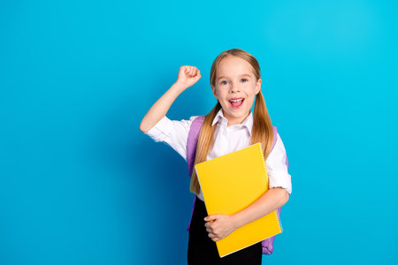 Smiling schoolgirl with ponytails holding a yellow notebook against a blue background, celebrating a...
