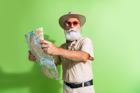 Photo of old serious professional old man hold map navigate for travel wear safari suit hat isolated green color backgroundの写真素材