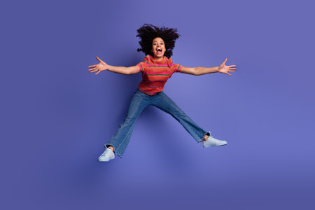 Excited young woman jumping in the air on a vibrant purple background wearing casual stylish clothes...