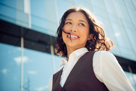 Confident businesswoman smiling outdoors with modern city glass building background on a sunny day.の写真素材