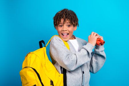 Excited young boy wearing grey cardigan with yellow backpack holding binoculars against a blue...