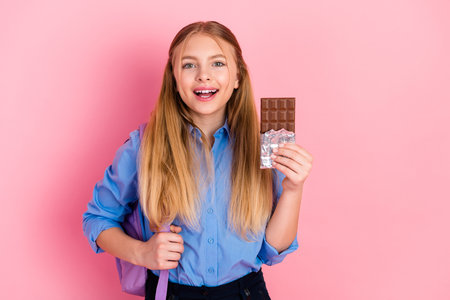 Cheerful preteen girl holding chocolate bar and backpack against pink background while wearing casual outfitの写真素材