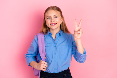 Smiling young schoolgirl with backpack making peace sign against pink background, celebrating...
