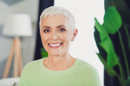 Smiling mature woman with short white hair in a casual sweater enjoying a sunny indoor setting...