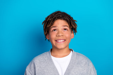 Happy schoolboy looking upwards against blue background, expressing curiosity and joy, wearing...