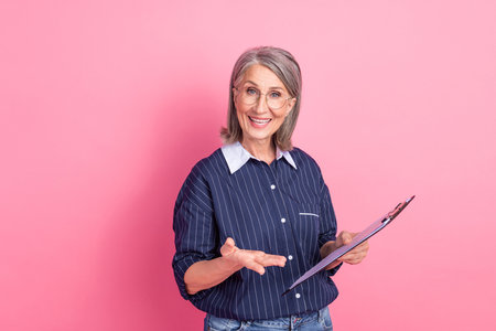 Confident mature businesswoman presenting with clipboard against vibrant pink background wearing stylish outfitの写真素材